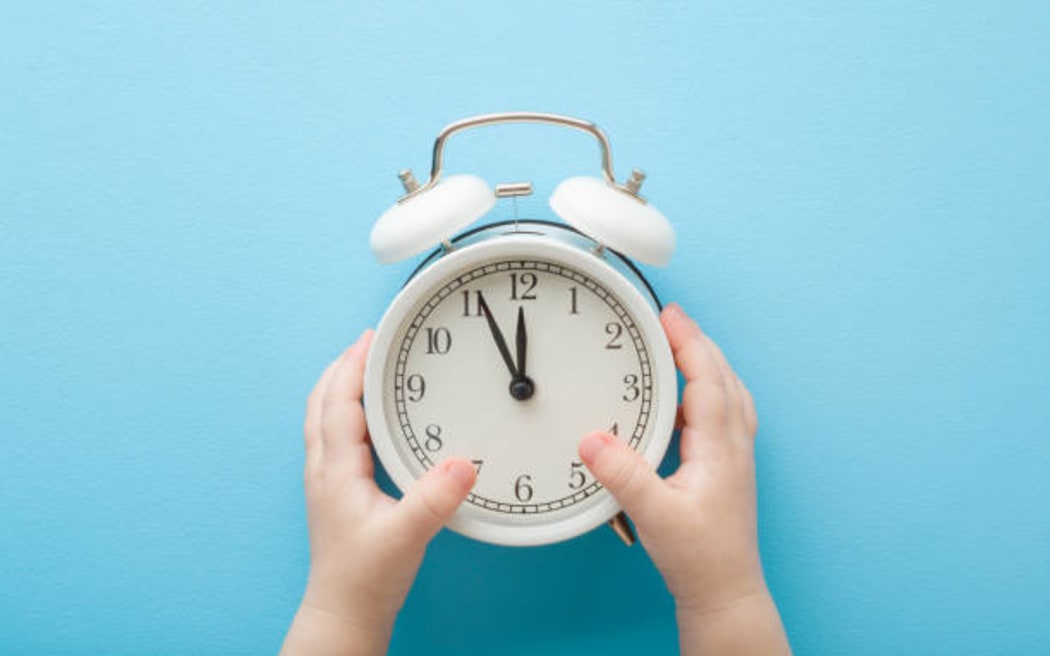 Baby hands holding white alarm clock on light blue table background. Pastel color. Time concept. Closeup. Point of view shot. Top down view.