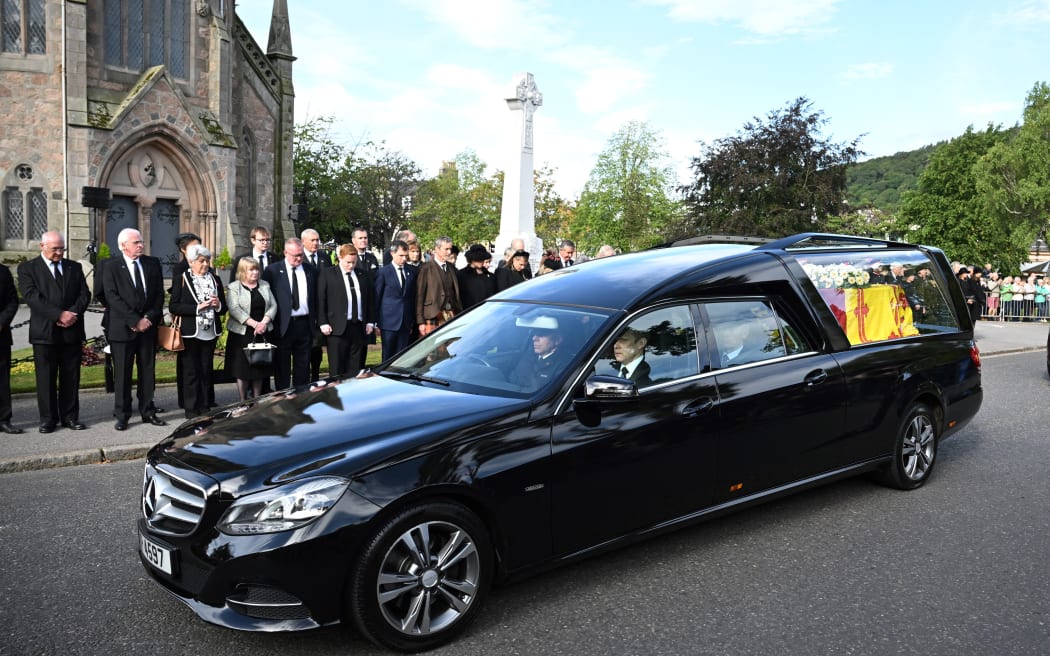 Members of the public pay their respects as the hearse carrying the coffin of Queen Elizabeth II, draped in the Royal Standard of Scotland, is driven through Ballater, on September 11, 2022. - Queen Elizabeth II's coffin will travel by road through Scottish towns and villages on Sunday as it begins its final journey from her beloved Scottish retreat of Balmoral. The Queen, who died on September 8, will be taken to the Palace of Holyroodhouse before lying at rest in St Giles' Cathedral, before travelling onwards to London for her funeral. (Photo by Paul ELLIS / AFP)