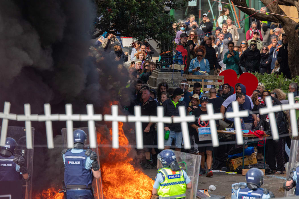 Police and occupiers face off across a fire under a garland of crosses