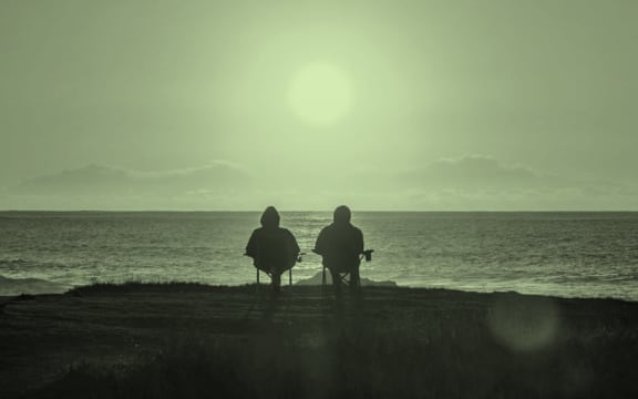 The image is of a pair of people in the foreground on a beach, watching the sun go down. Both are sitting on folding chairs.