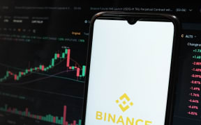 A Binance logo is seen displayed on a smartphone screen and an Exchange graph on a MacBook screen.