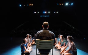 A man sits in a chair on a stage with his back to the camera. Children surround him in a U-shape on the floor.
