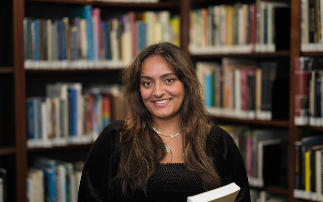 Anjuli smiles at the camera. She is standing in a library, holding a book. She wears a black top.