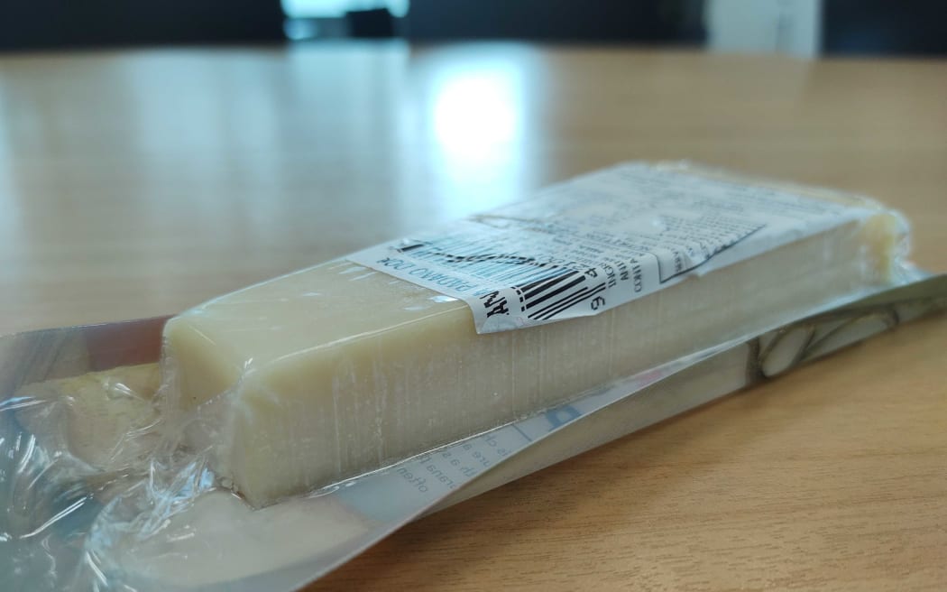 A slice of cheese on a table in the RNZ kitchen. It is clearly a hard cheese, with the cut side visible and a slice of rind in the background.