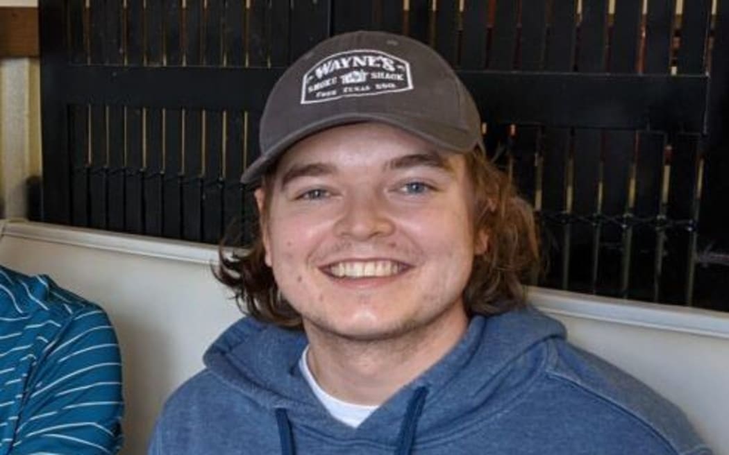 Christian Glass who has shoulder length brown hair is wearing a blue hoodie, a cap and is looking directly at the camera and smiling.