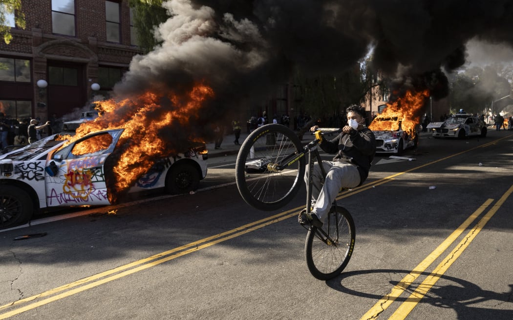 A demonstrator rides a bicycle past burning Waymo vehicles as protesters clash with law enforcement near the federal building during a protest in response to federal immigration operations in Los Angeles, California, on June 8, 2025. US President Donald Trump deployed 2,000 troops on June 7 to handle escalating protests against immigration enforcement raids in the Los Angeles area, a move the state's governor termed "purposefully inflammatory." Federal agents clashed with angry crowds in a Los Angeles suburb as protests stretched into a second night Saturday, shooting flash-bang grenades and shutting part of a freeway amid raids on undocumented migrants, reports said. (Photo by ETIENNE LAURENT / AFP)
