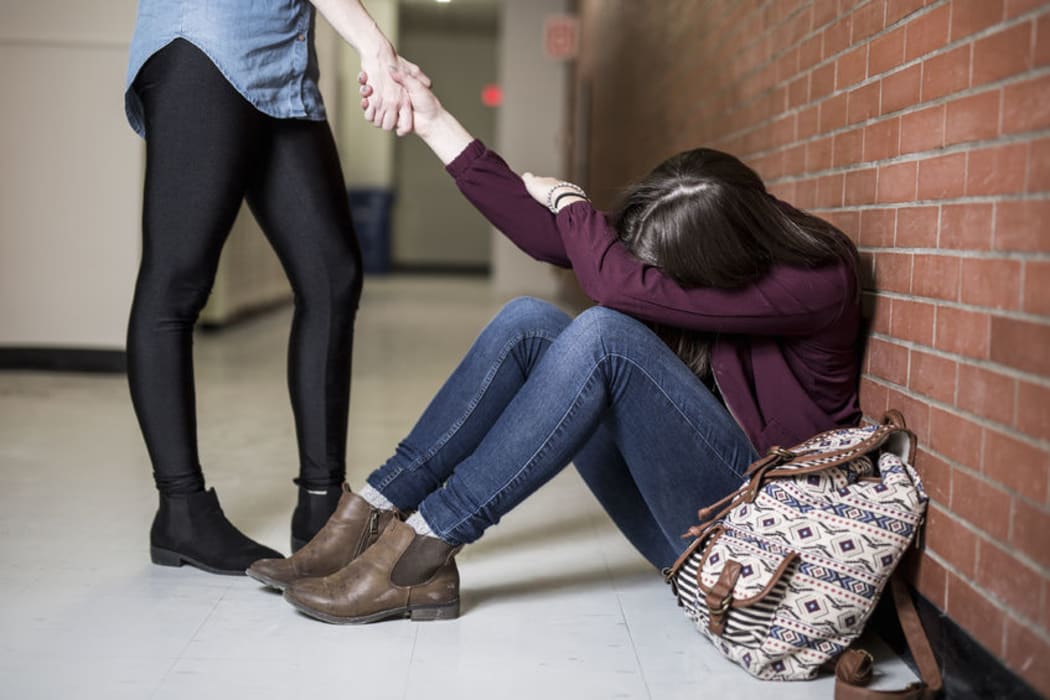 A photo of a teenager trying to pull her anxious friend up from the floor in a school corridor