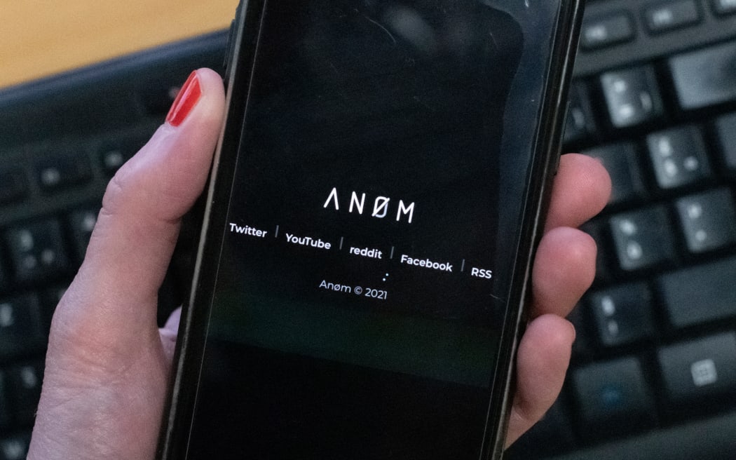 An illustration picture shows the AN0M logo displayed on the screen of a smartphone.
