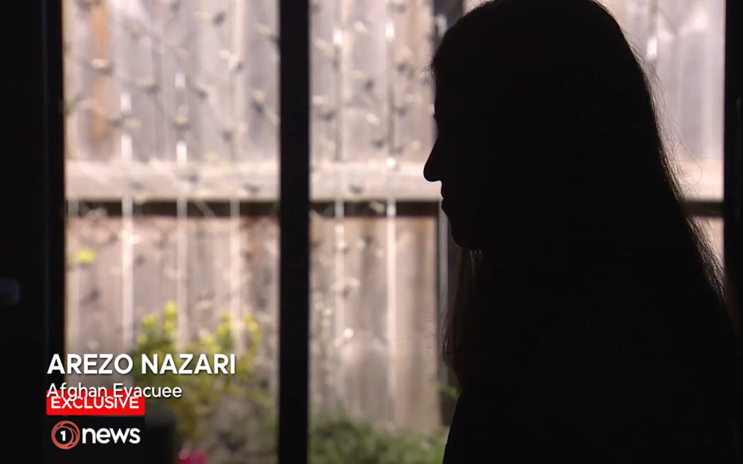 A screenshot of a TV news segment. A girl is sitting indoors in profile, backlit by a lounge window. She is in darkness, so you cannot see her face. Below, text reads "AREZO NAZARI - AFGHAN EVACUEE"