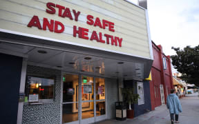 A pedestrian walks past a shuttered movie theater, with the message 'Stay Safe and Healthy' displayed on the marquee.