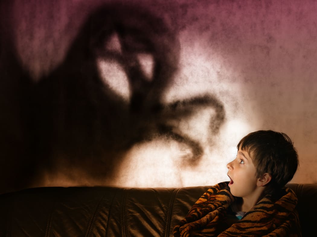 A photo of a boy in the dark with a shadow behind him in the shape of a monster with its arms extended.
