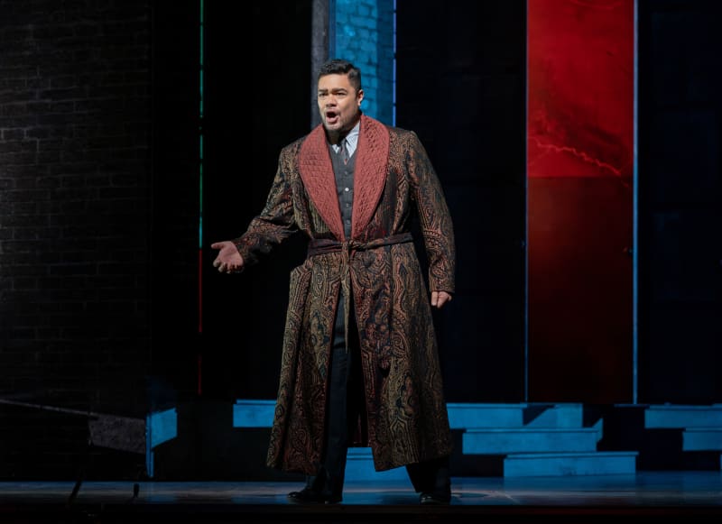Pene stands on stage in a rich paisley robe draped over a suit. He is singing and gesturing.