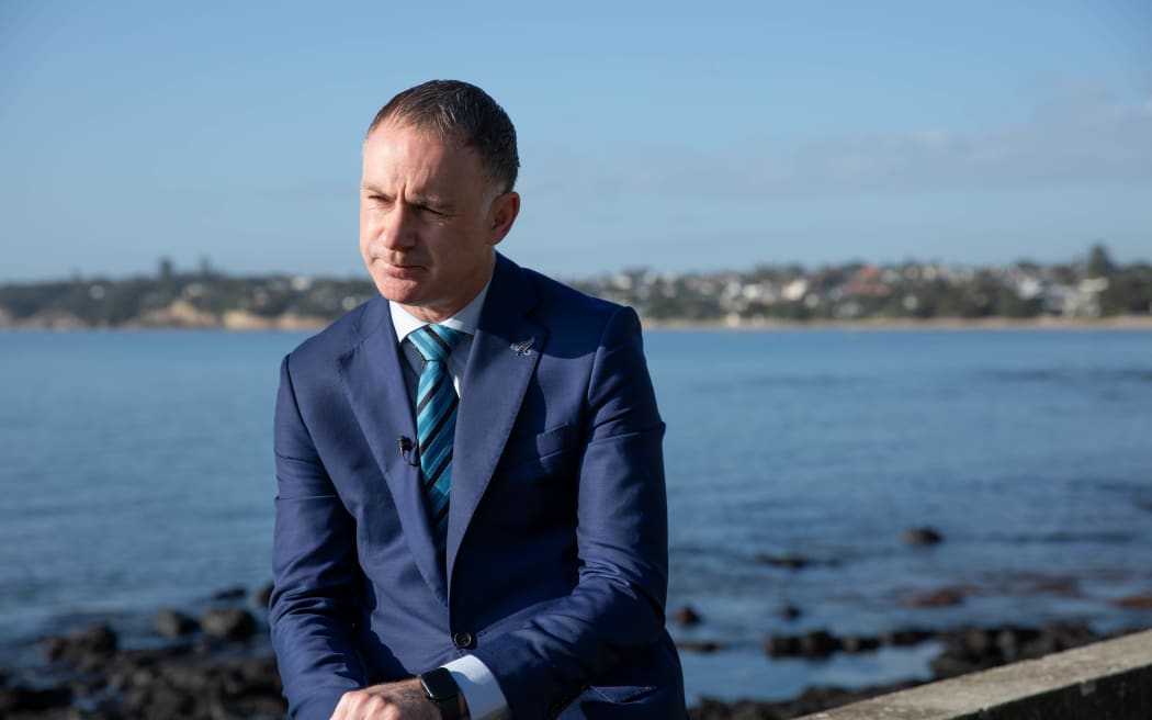 Climate Change minister Simon Watts, wearing a blue suit, sits on a seawall on Auckland's North Shore.