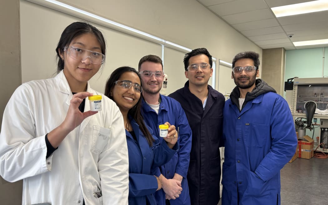 A group portrait shot of 5 people in lab coats, wearing safety glasses. Two are holding small specimen containers.