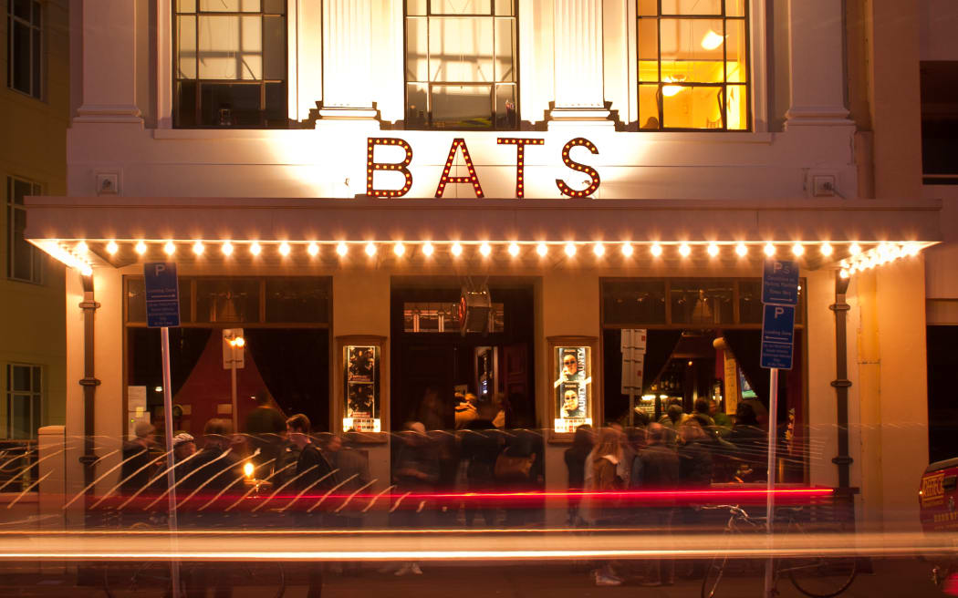 The BATS Theatre building from the streetside at nighttime. The old building facade is lit up in golden light. The word BATS is written in lights. People are inside having a good time.