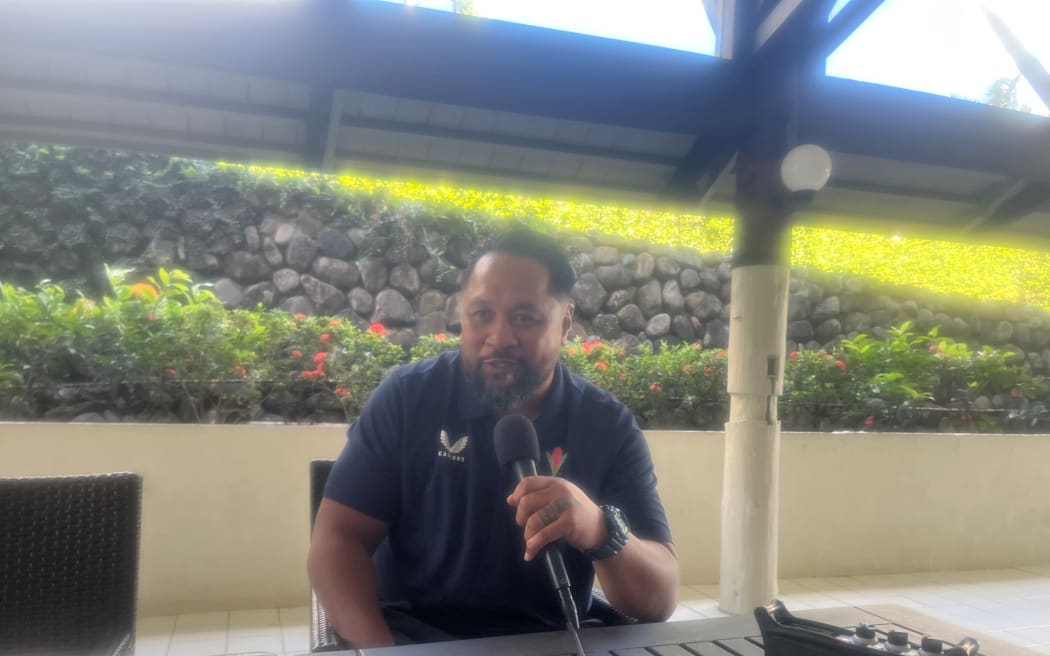 A photo of Vaovasamanaia Seilala Mapasua, the head coach of Manu Samoa sitting outside at a table holdig a microphone