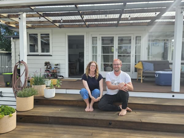 Two friends, a woman and a man are sitting side by side on the outside steps of a deck, smiling at the camera. The deck has a roof and in the background you can see pots of flowers, a couch and a chilli bin