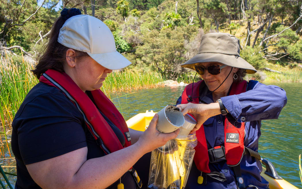 Two women wearing red lifejackets pour mud out of PVC pipes into ziploc plastic bags while sitting on a boat in the middle of a lake fringed by raupō reeds and bush.