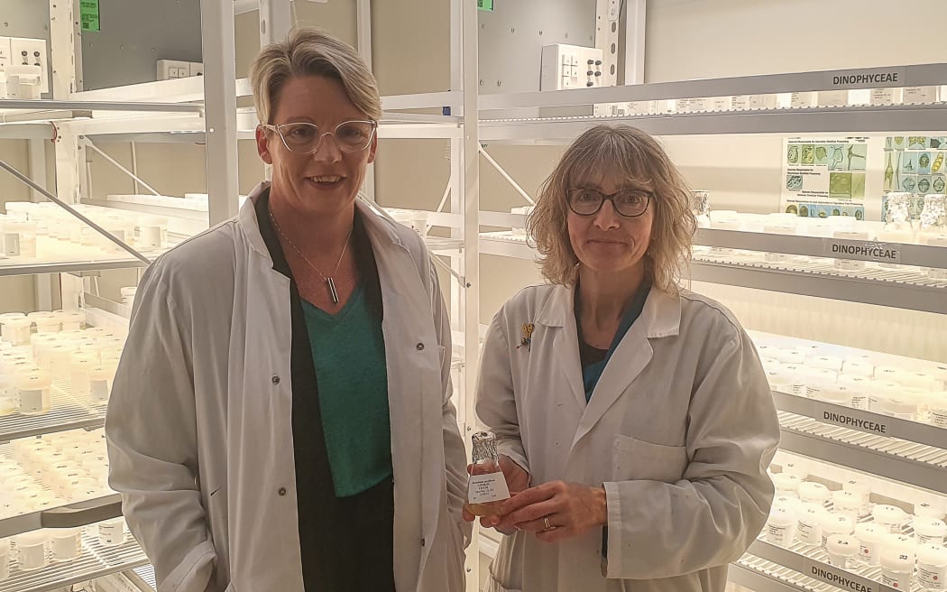 Two women in white labcoats standing in front of racks of vials and flasks. The womean in the right is holding a conical flask.
