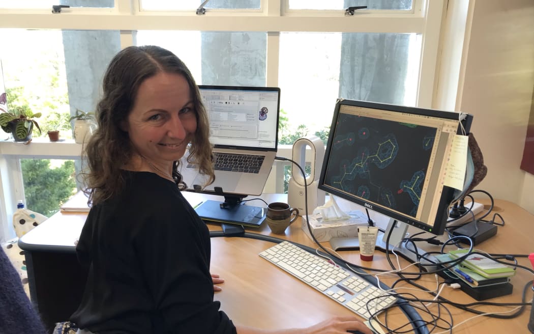 Adele sits at her office desk in front of her computer. There's a protein structure on the screen in front of her, she is looking towards the camera.