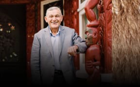 Kiingi Tuheitia Pootatau Te Wherowhero VII, the Māori King stands outside a Whare Nui smiling in a grey suit. The image is frames with a detail of a feather korowai.