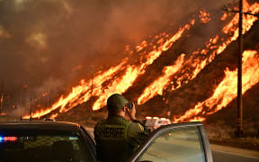 A sheriff takes photos as flames from the Hughes Fire race up the hill in Castaic, a northwestern neighborhood of Los Angeles, California, on January 22, 2025. A new wildfire erupted north of Los Angeles on January 22, exploding in size and sparking thousands of evacuation orders in a region already staggering from the effects of huge blazes.
Ferocious flames were devouring hillsides near Castaic Lake, spreading rapidly to cover 5,000 acres (2,000 hectares) in just over two hours.
The fire was being fanned by strong, dry Santa Ana winds that were racing through the area, pushing a vast pall of smoke and embers ahead of the flames.
Evacuations were ordered for 19,000 people all around the lake, which sits around 35 miles north of Los Angeles, and close to the city of Santa Clarita. (Photo by Frederic J. Brown / AFP)