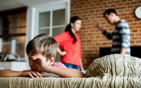 Parenting arguing in background with child wiping tears on sofa.