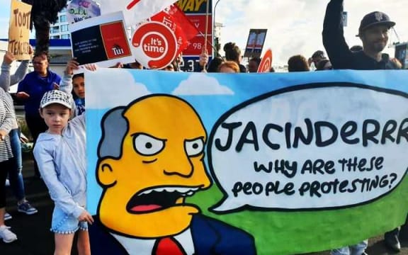 child holding large banner with cartoon head and speech bubble saying: "Jacindaa why are these people protesting?"