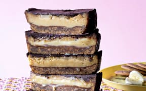 A vertical stack of five pieces of banana caramel slice, showing a cross-section of biscuit base, gooey caramel and chocolate topping, on a pink patterned cloth.