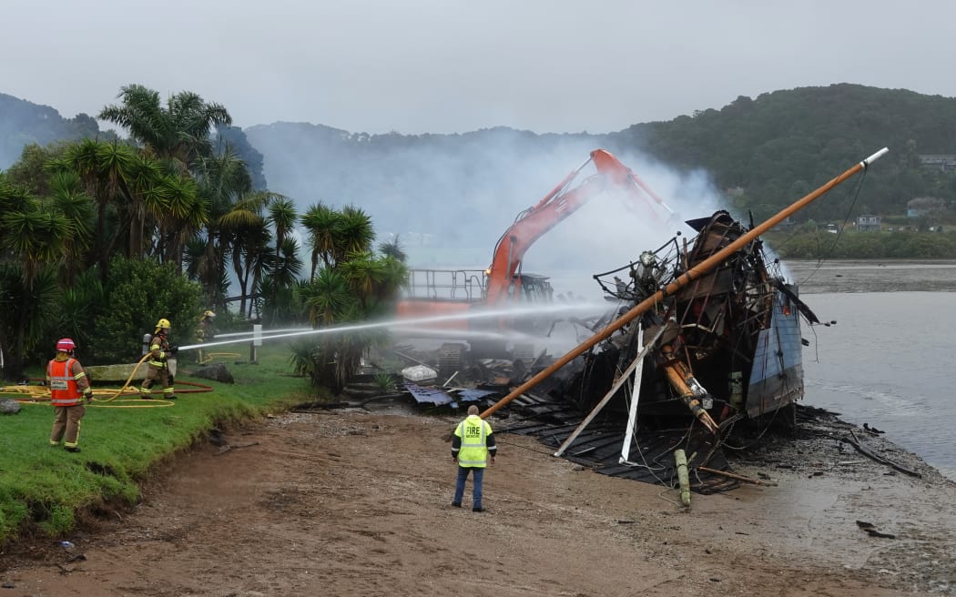 A digger demolishes what's left of the Tui as firefighters continue dampening down the wreckage.