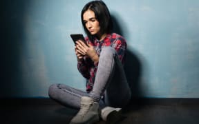 A photo of a young girl sitting in the dark gazing at her phone.