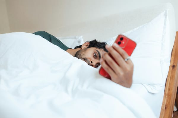 A man in bed alone looks at a red phone.