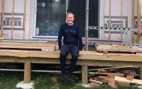 Chris Bennett sits on the deck of an unfinished house. He is wearing a navy long sleeved shirt and dark coloured pants and shoes. He smiles at the camera. The house is covered in scaffolding.