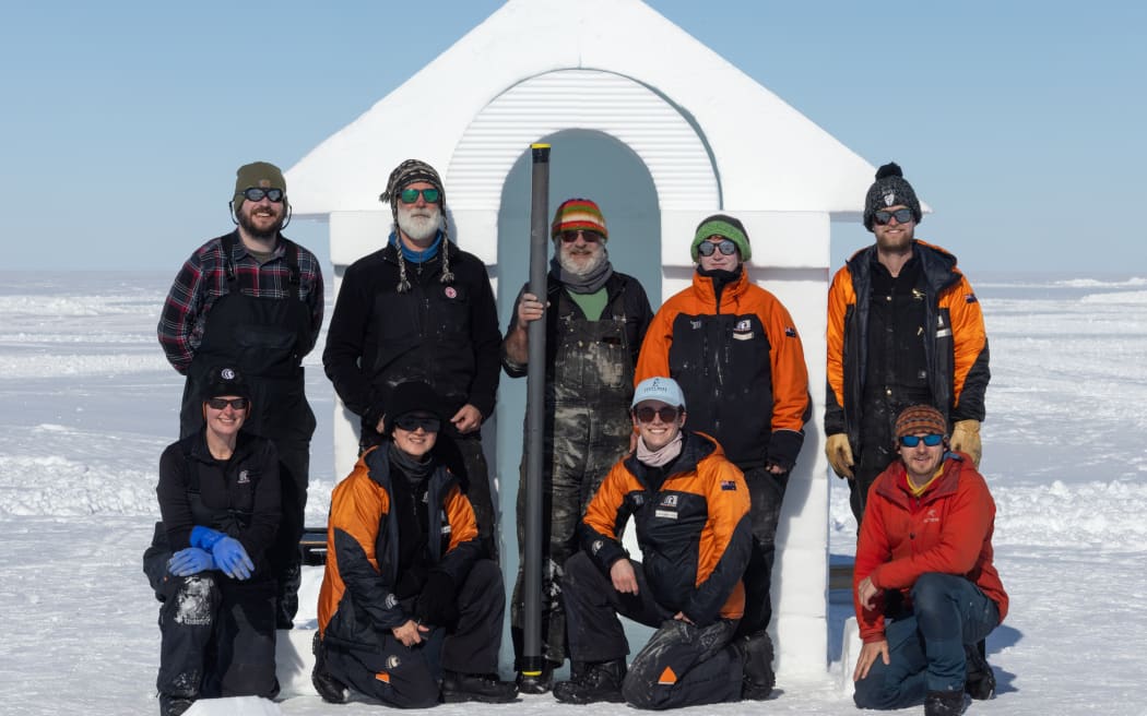 Nine people in two rows wearing warm clothes, sunglasses and beanies stand in front of a small white hut on the ice beneath a blue sky. The man in the middle is holding a long cylinder that is taller than him.