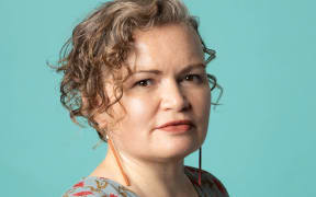 Tina stands in front of a blue background. She has cheek length wavy hair falling dow the left hand side of her face. She is wearing earrings which are like long orange, dangly sticks.