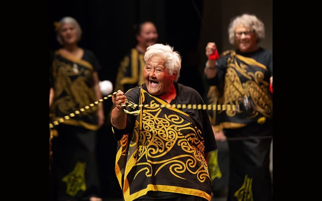 A kuia twirls a poi on stage with her group. She smiles widely.