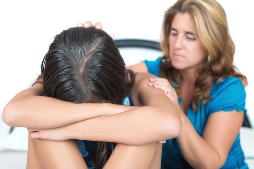 A photo of a mother comforting her upset teenage daughter who is sitting with her head on her hands