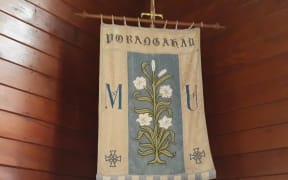 A fabric banner with a stem of white lilies sewn on is displayed in a corner at the front of St Michael and All Angels Church, Porangahau.