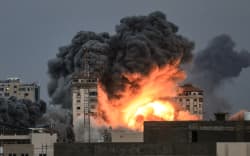 People standing on a rooftop (left), watch as a ball of fire and smoke rises above a building in Gaza City on 7 October, 2023 during an Israeli air strike.