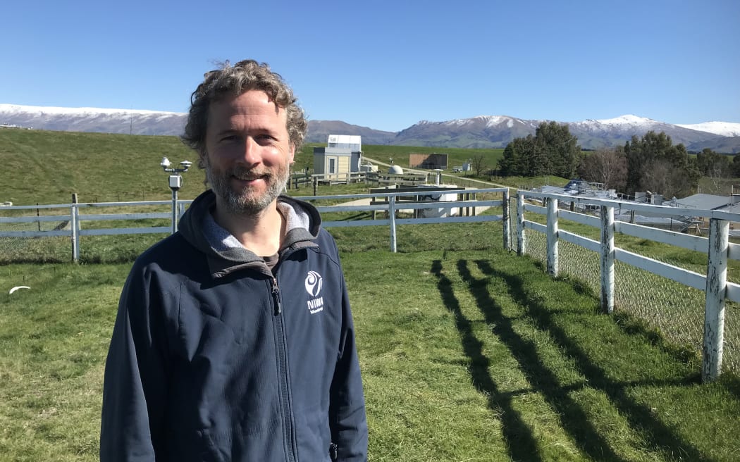 A man wearing a NIWA-banded navy fleece stands in a grassy paddock with a small building in the background. Further in the background are snow-capped mountains beneath a bluebird sky.