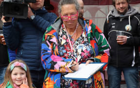 Julia stands in a crowd holding a clipboard and watching a game of marbles. She is wearing pink spectacles and wearing a multi-coloured jacket.