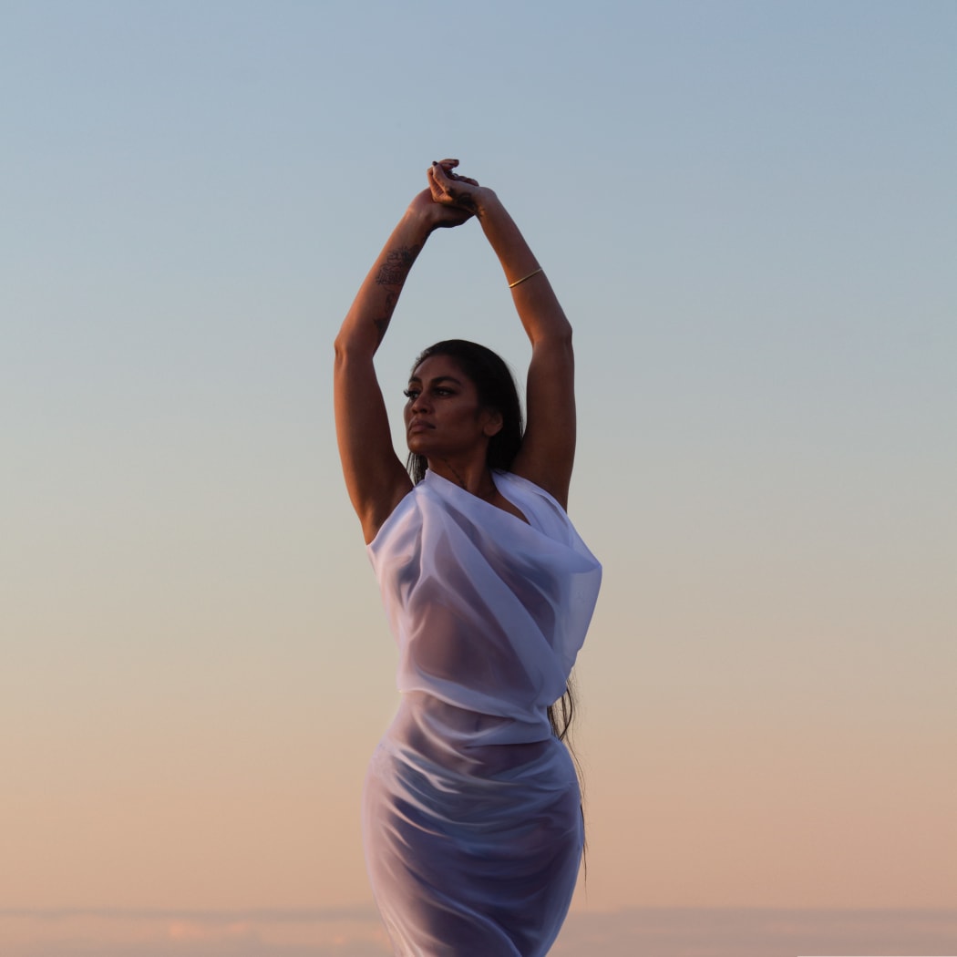 The album cover for 'Sweet Surrender'. Singer/songwriter Aaradhna poses against the background of the setting sun at the beach. Her arms in the air.