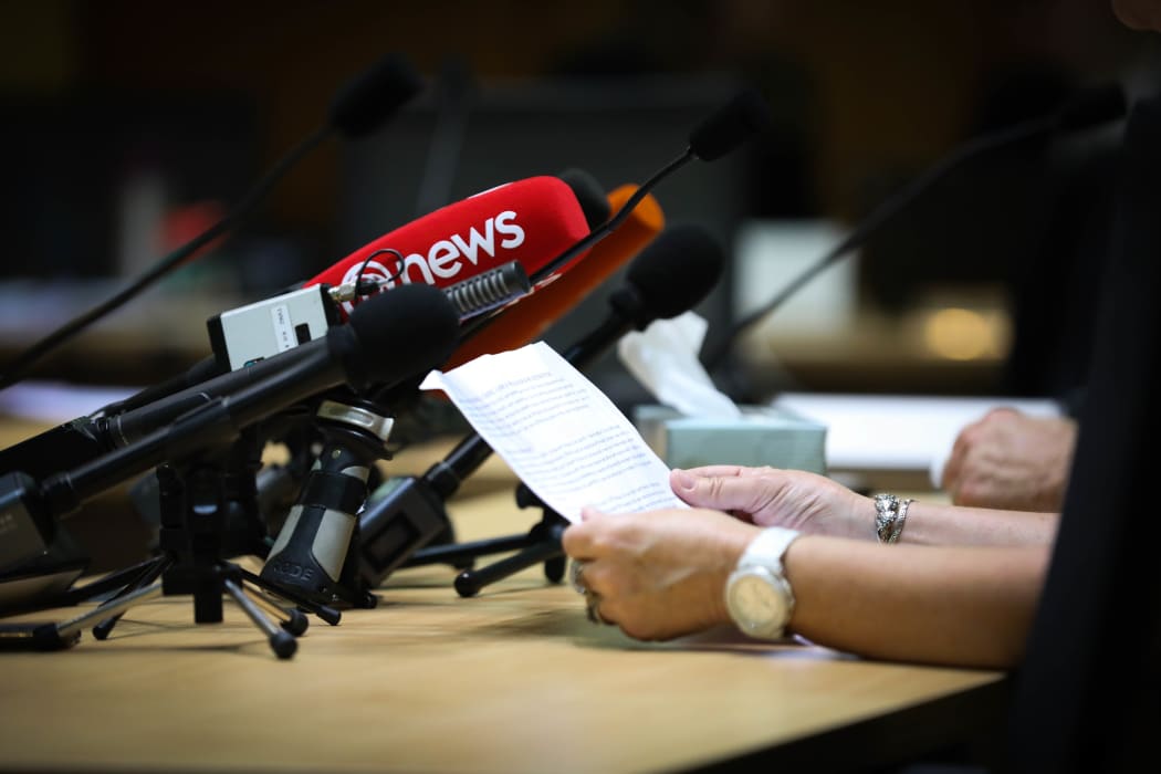 A submitter reads their written submission to a select committee and a lot of media microphones. Tissues on the table indicate the topic is sensitive.