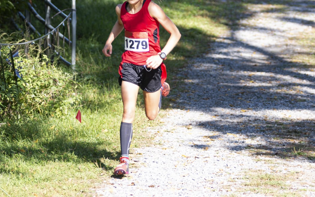 A female high school runner is in the lead during a cross country race running downhill on a gravel path on a bright sunny day.