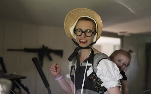 Simone Collins, wearing a pilgrim-style dress topped off with a bonnet, carrying one of her children on her back