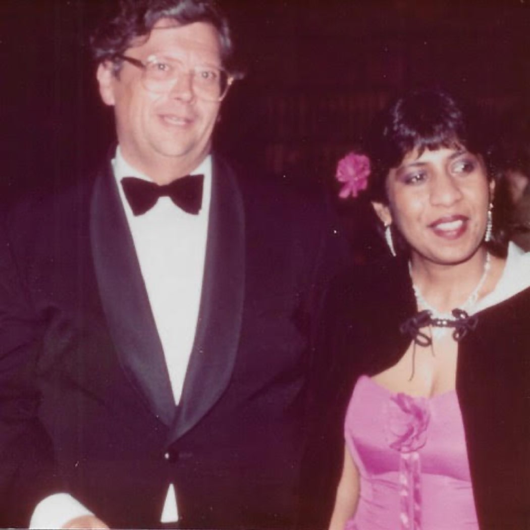 David and Jeya stand side by side, smiling. They are both in formalwear, David in a black suit jacket and black bowtie, and Jeya in a pink dress with a black cape over the top.