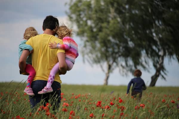 A man in a yellow t-shirt holds two blonde children in a field.