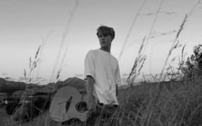 Max stands in a field holding a guitar and gazing wistfully into the distance.