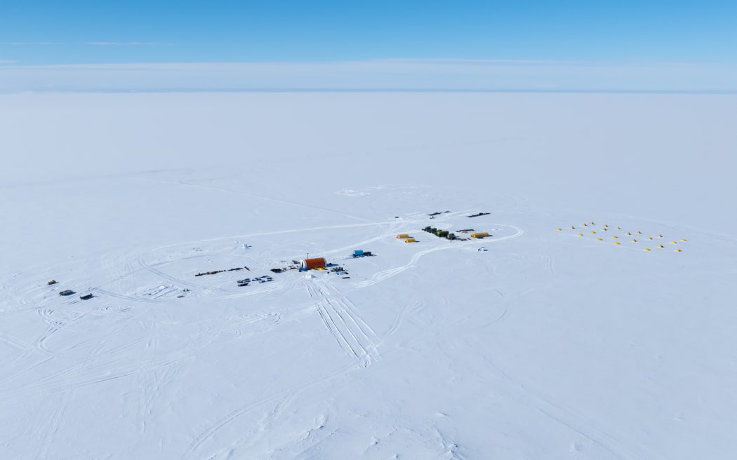 A view of an Antarctic field camp, made up of tents and modified shipping containers, viewed from above with the white of the continent stretching away to the horizon.