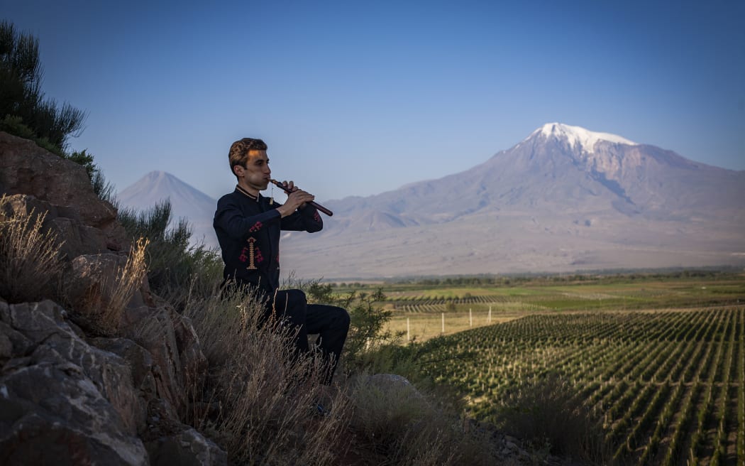 Armenian duduk player Arsen Petrosyan plays the instrument sitting outside, with a snow-capped mountain in the background.